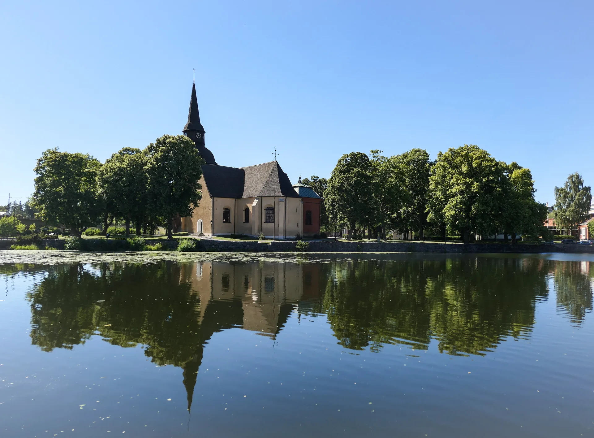 En ljus stenkyrka med spetsigt torn står bland höga lövträd vid vattnet i Eskilstuna en klar sommardag