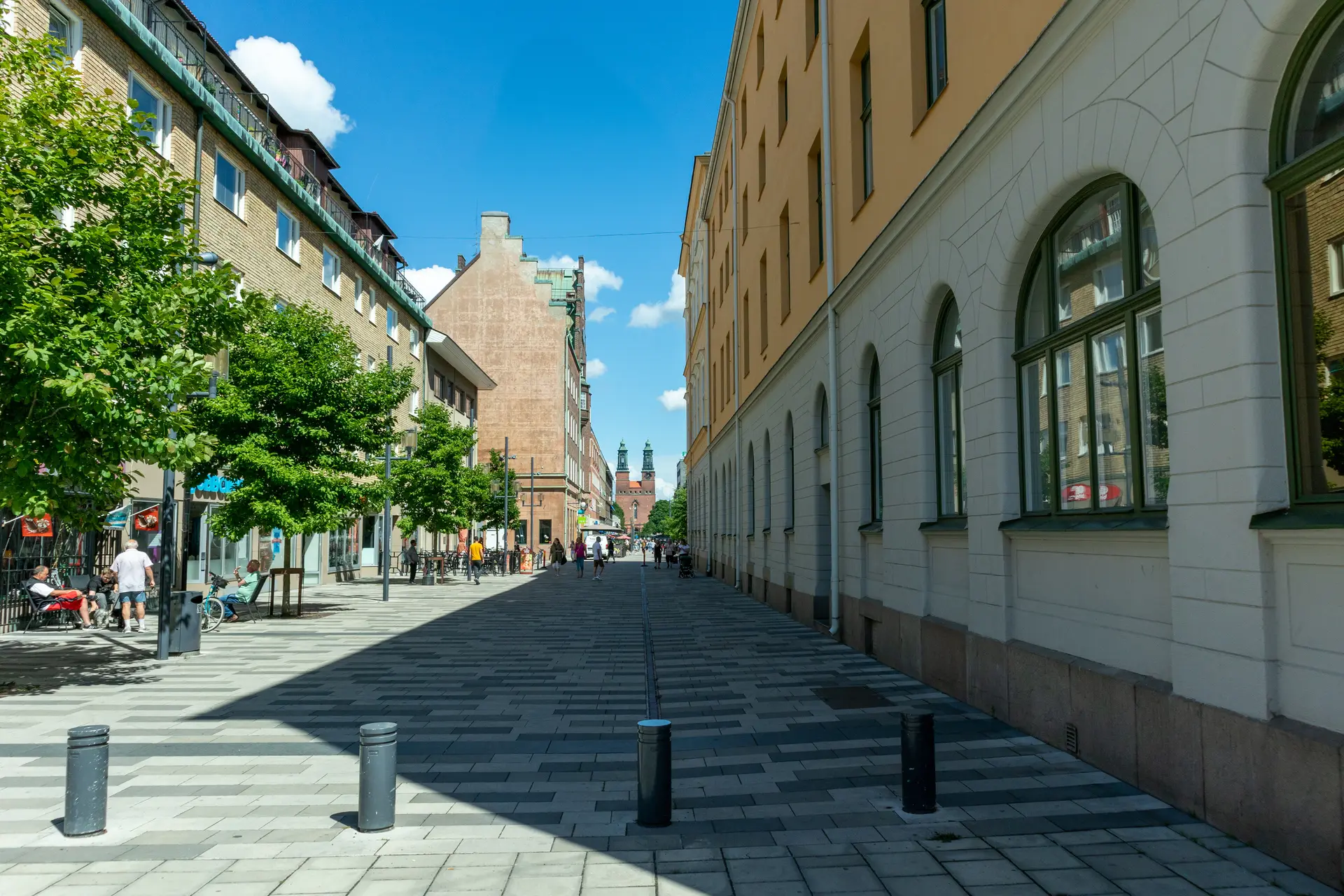 Sommardag på en bilfri gata i centrala Eskilstuna, med lövträd längs husraden och två kyrktorn långt bort i fonden