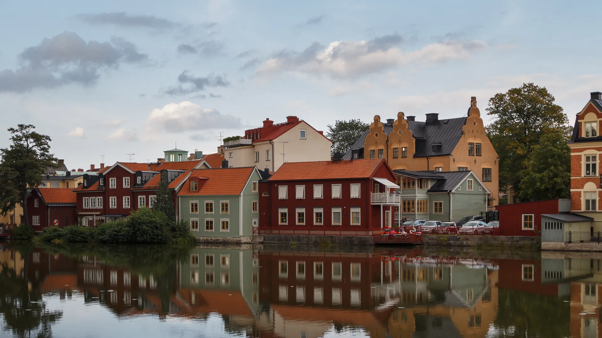 Röda och ljusgröna trähus vid vattnet speglar sig i den stilla ytan en molnig kväll i centrala Eskilstuna